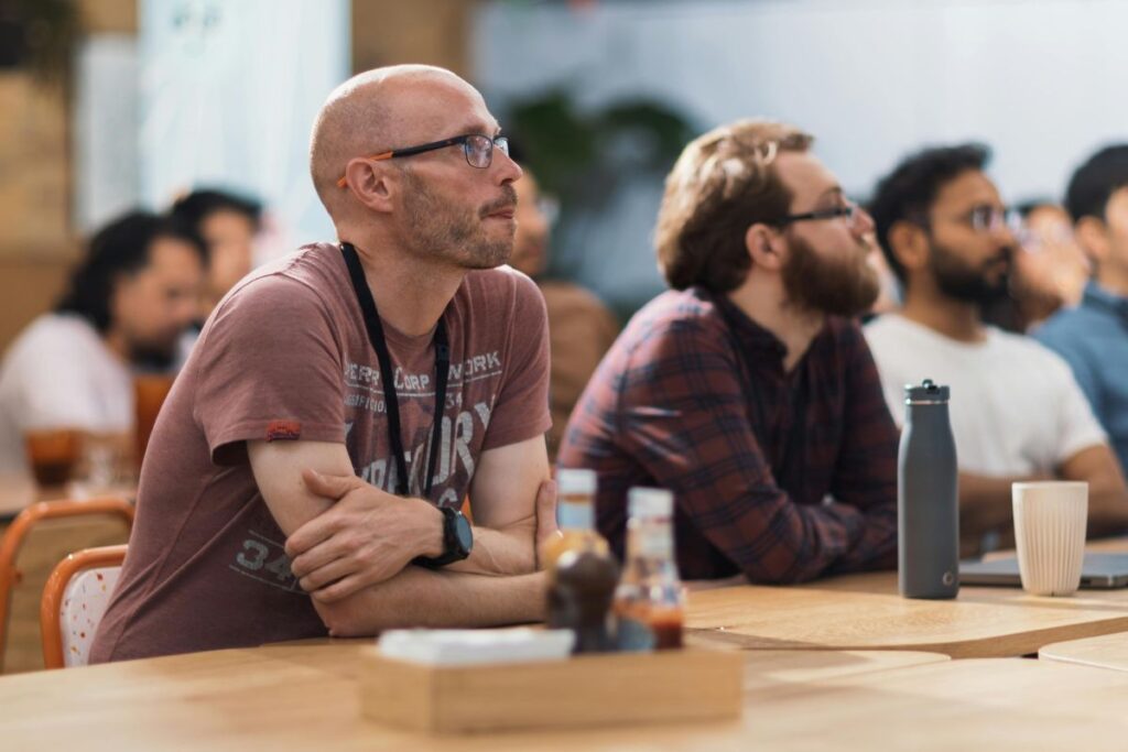A man with glasses sits in a work conference and listens to the speaker.