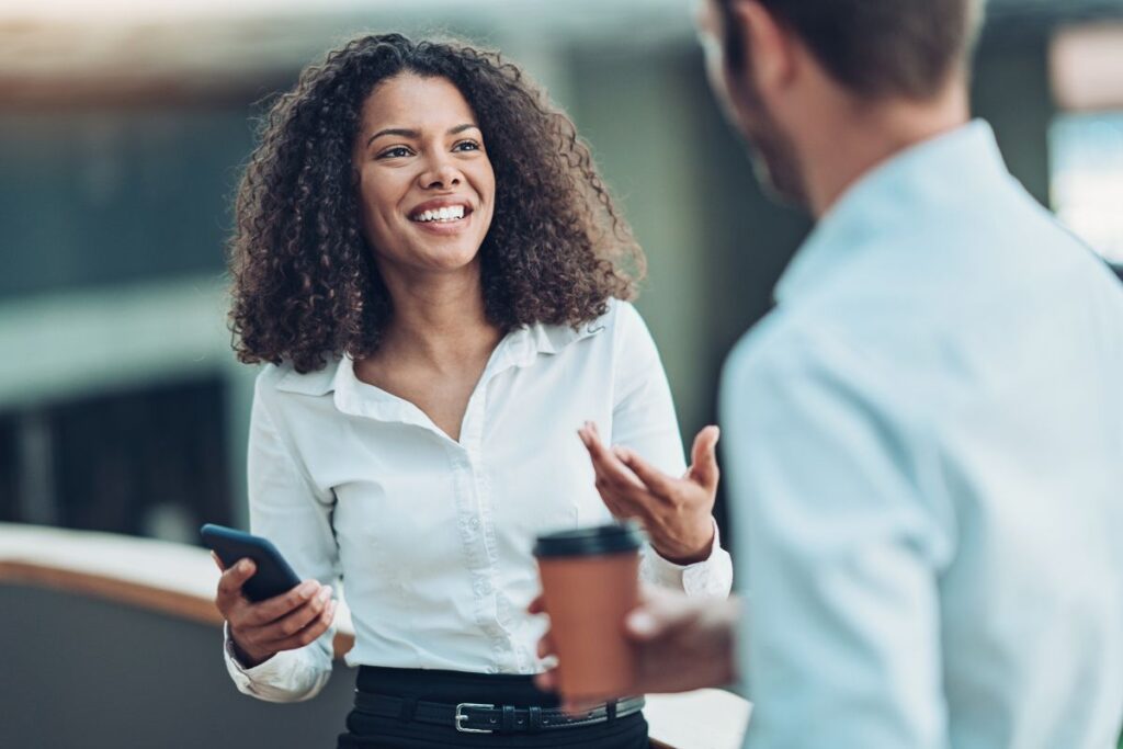 A woman chats to her co-worker. She is holding her phone and he is holding a coffee.