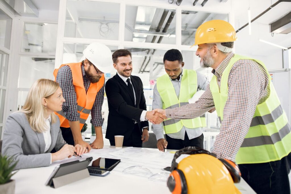 A man in a black suit shakes hands with a man in a hardhat and a hi-vis jacket. There are two other men stood up in the meeting, and one woman in a suit who is sat down.