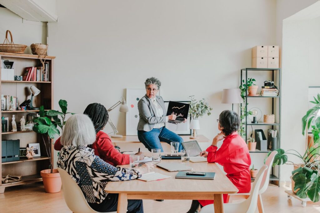 A senior leadership team has a meeting. They are a group of four women. One woman sits in front and presents a graph to the group.