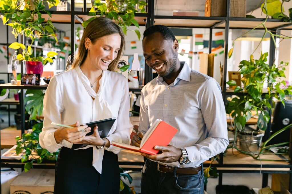 Two employees stand together and smile. They are both looking in the same notebook.