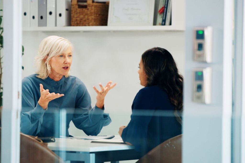 Two women sit in a meeting together. The woman leading the meeting is gesturing with her hands.