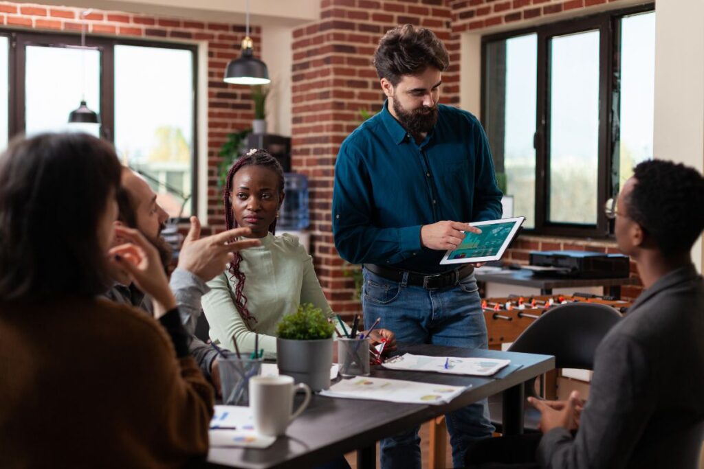 A group of co-workers sit in a meeting together and discuss data. One man stands up and presents graphs on a tablet.