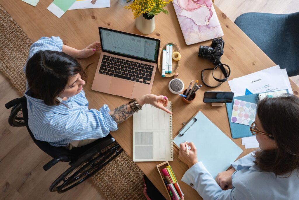 a birds eye view perspective of two women working together. One woman sits in front of a laptop, the other writes in a notepad. One of the women is in a wheelchair.