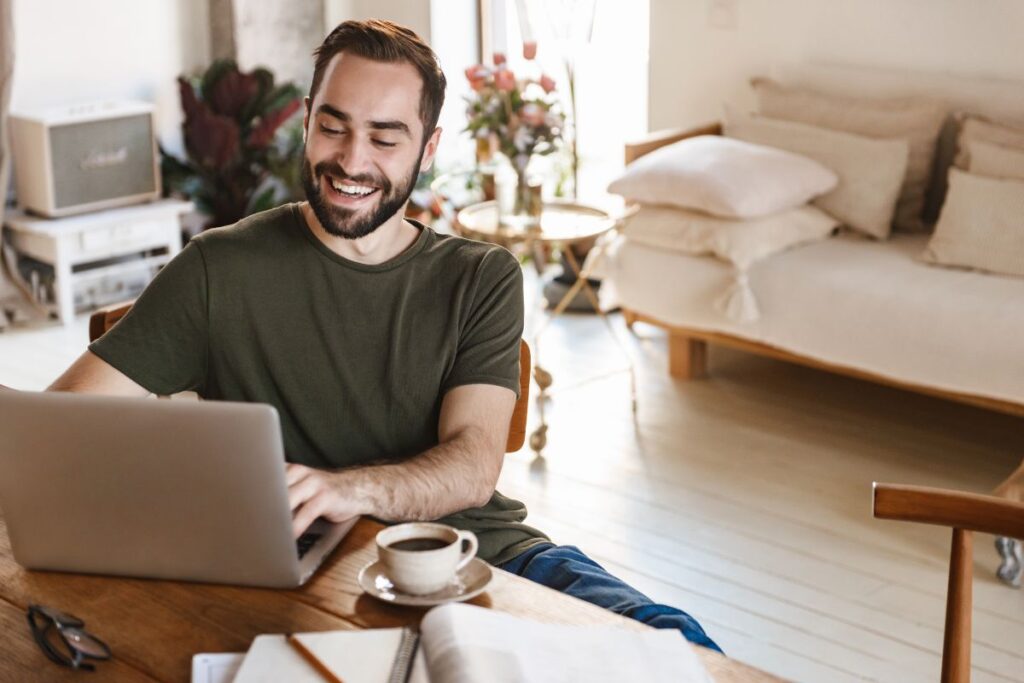 A man sits in front of his laptop and works from home. He is smiling and there is a cup of coffee in front of him.