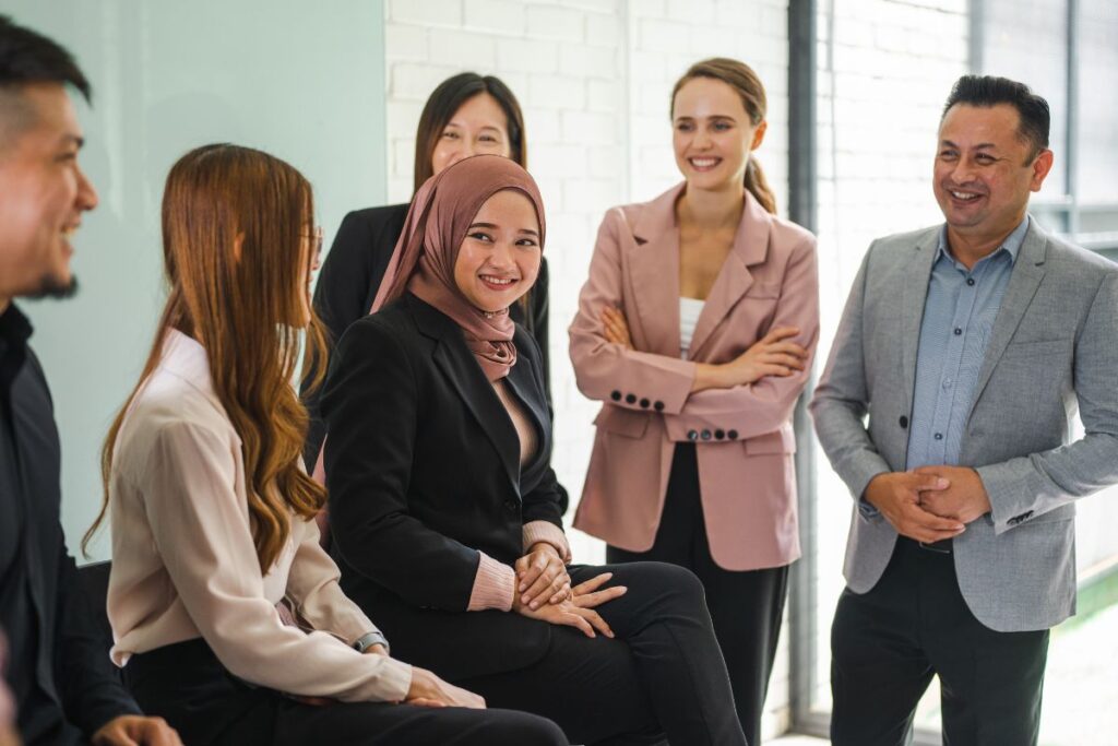 A diverse group of employees sit together, smiling and laughing. They are all dressed in professional clothing.