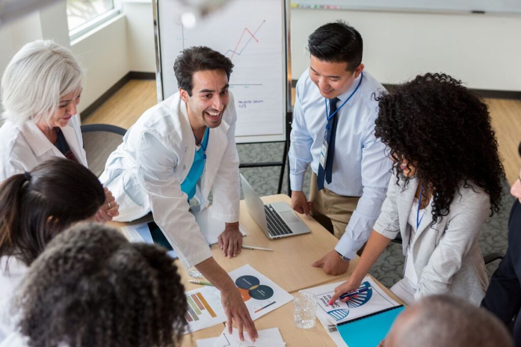 A group of hospital and healthcare workers in white coats and blue scrubs present graphs to people in suits. They are all smiling.