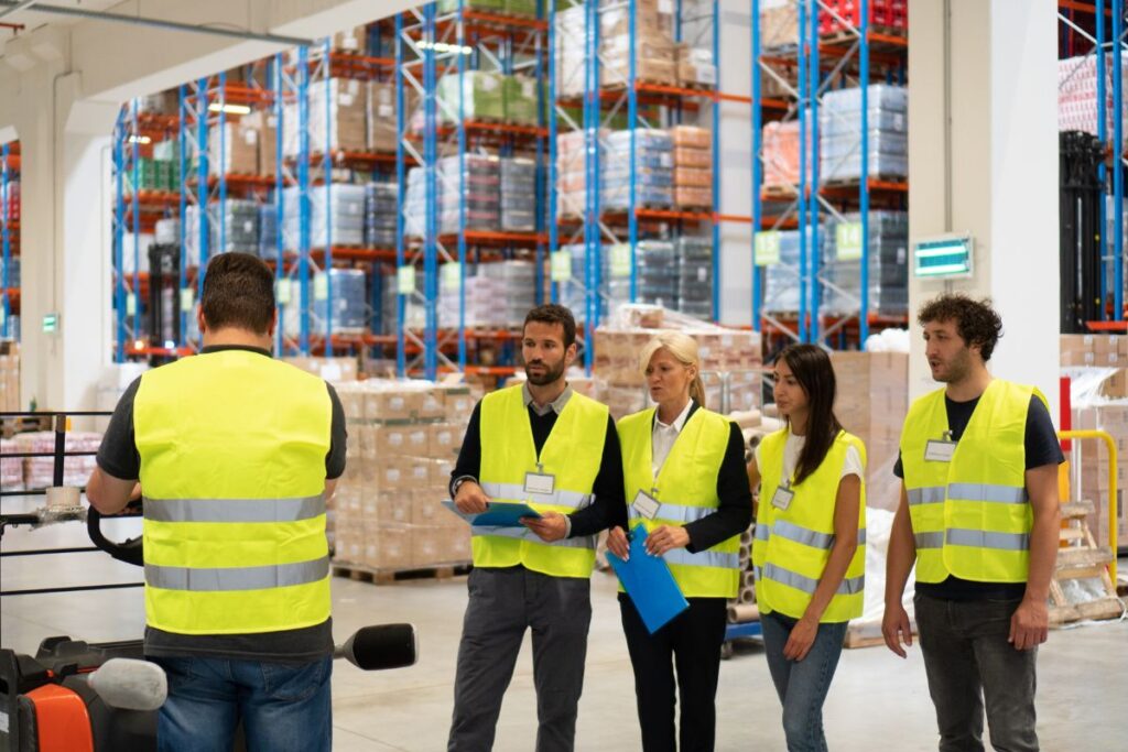 A group of professionals take management training in a warehouse setting. They are all wearing hi-vis jackets.