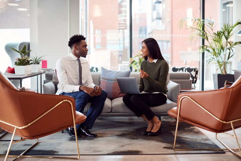 A woman and a man sit facing each other on a sofa. The woman conducts an interview with the man. She has a laptop on her lap.