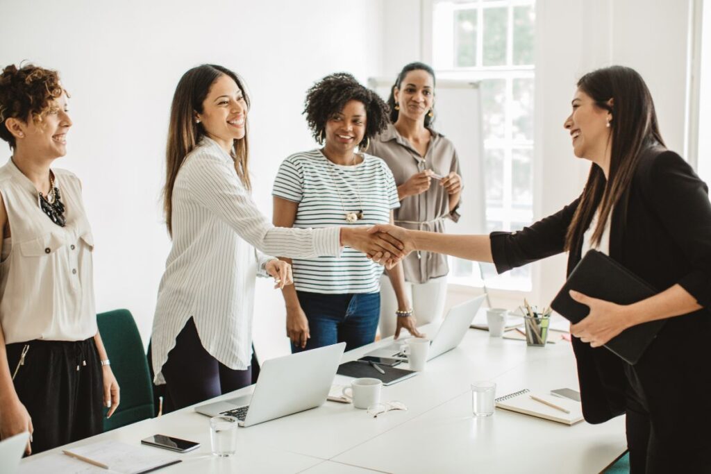 Four women interviewers stand on one side of a table. A woman stands on the opposite side and shakes hands with one of the interviewers. They are all smiling.