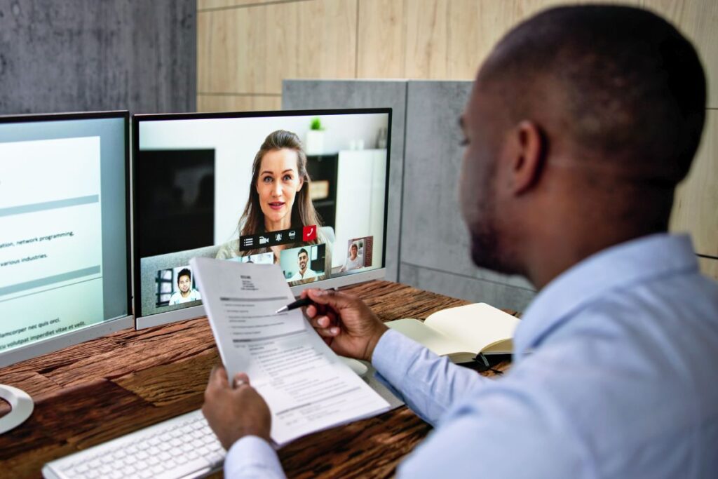A man conducts an interview online. He is talking to a woman over a video call. He is holding candidate information.