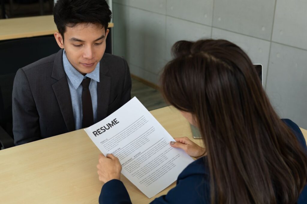 A woman sits in front of a man and interviews him. She is holding his resume.