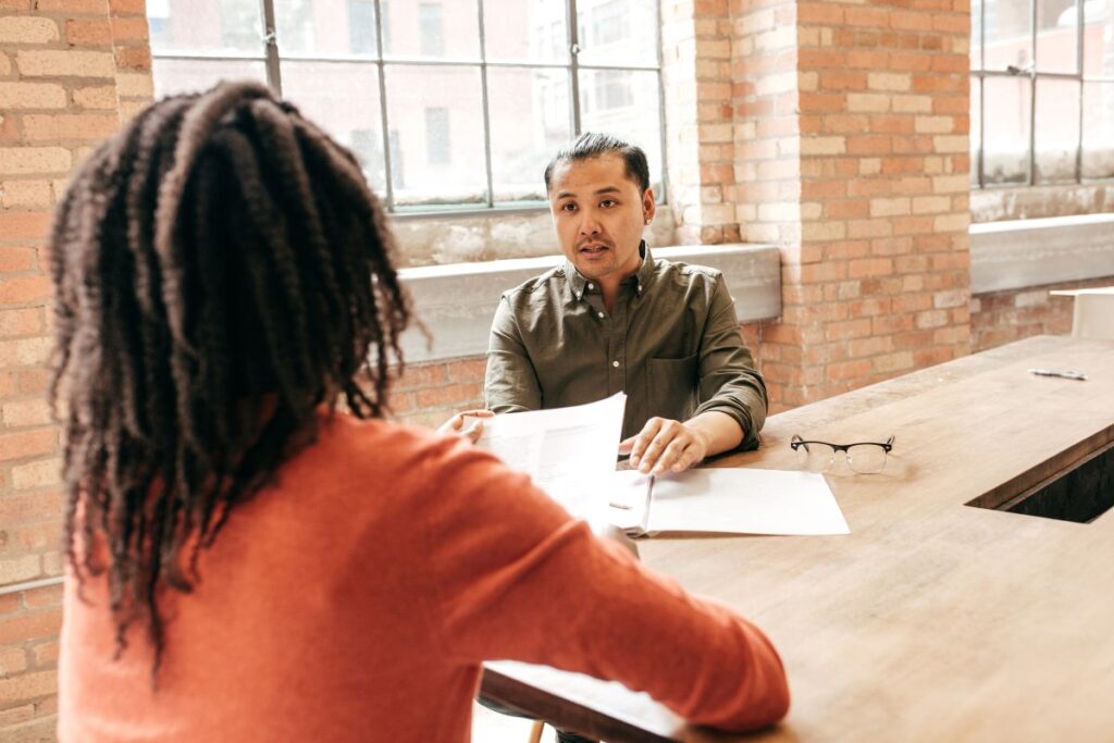 A woman conducts an interview with a man. She is holding his resume. He is answering a question. His glasses are next to him on the table.