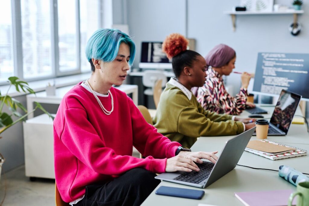 A man with blue hair sits at his desk in work. He is sat at the forefront of two women who are also working.