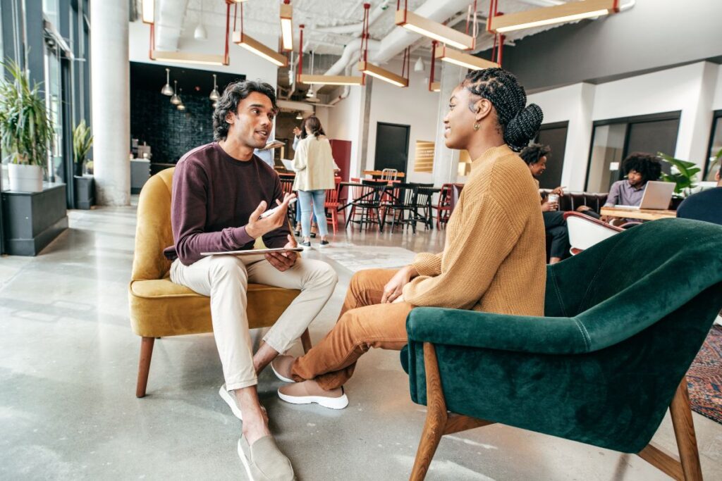 A man conducts a casual interview with a woman. They are both sat on armchairs in a shared working space.