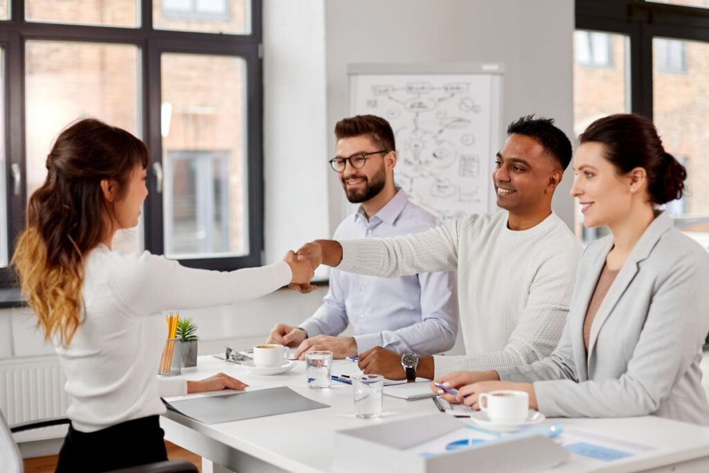Two men and one woman sit on one side of a table. A second woman sits on the other side of the table. She shakes hands with the man in the middle. They are all smiling.