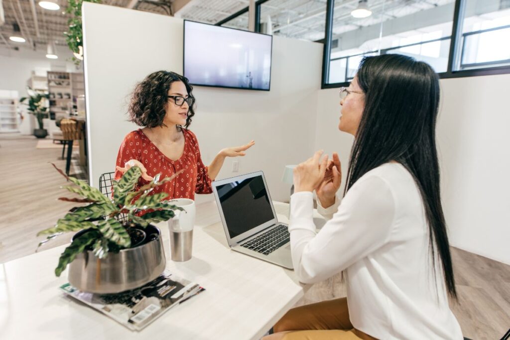 Two women sit across from each other in an interview setting. One woman has a laptop in front of her. They are both talking and smiling.