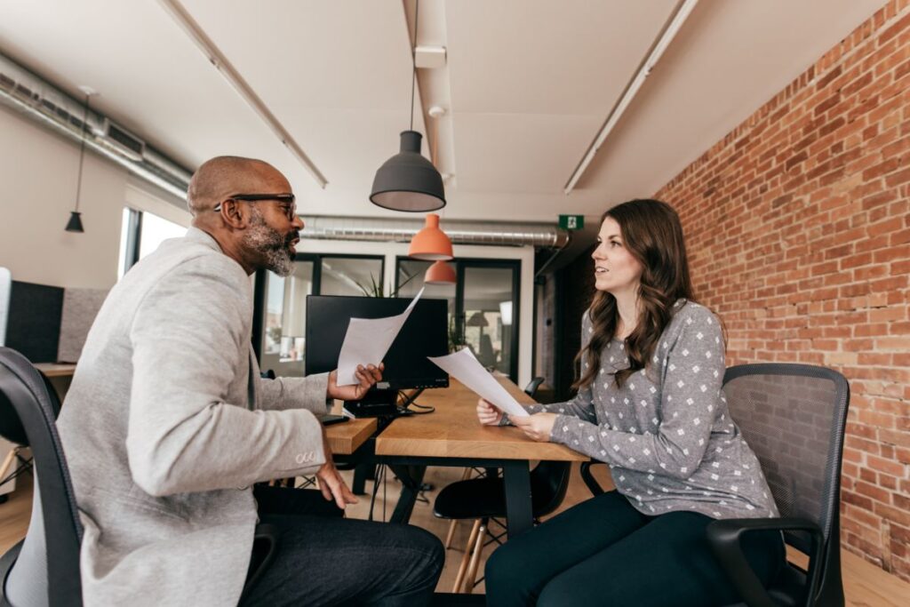 A woman and man sit opposite each other in an interview setting. They each hold note papers.
