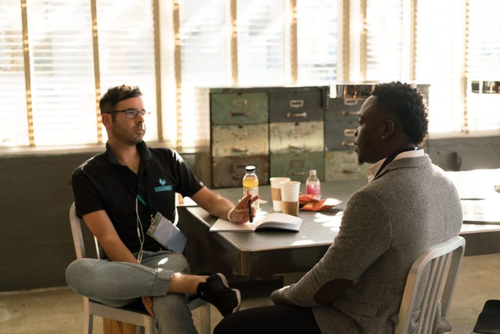 Two men sit opposite each other in an interview setting. One holds a pen and rests his hand on a notebook.