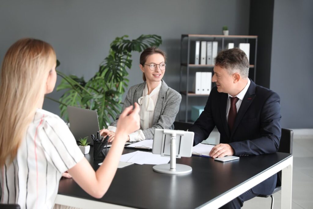 A man and a woman sit opposite a woman in an interview setting. There is a laptop and notes in the desk in front of them. They are smiling.