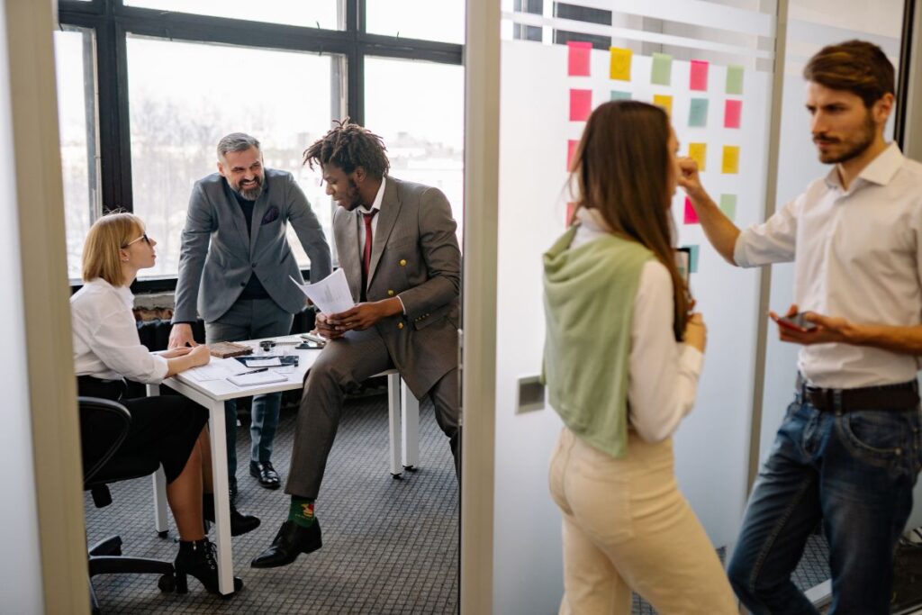 Two employees stand outside of a meeting room and chat. Within the meeting room, three members of staff chat to each other in a meeting setting.