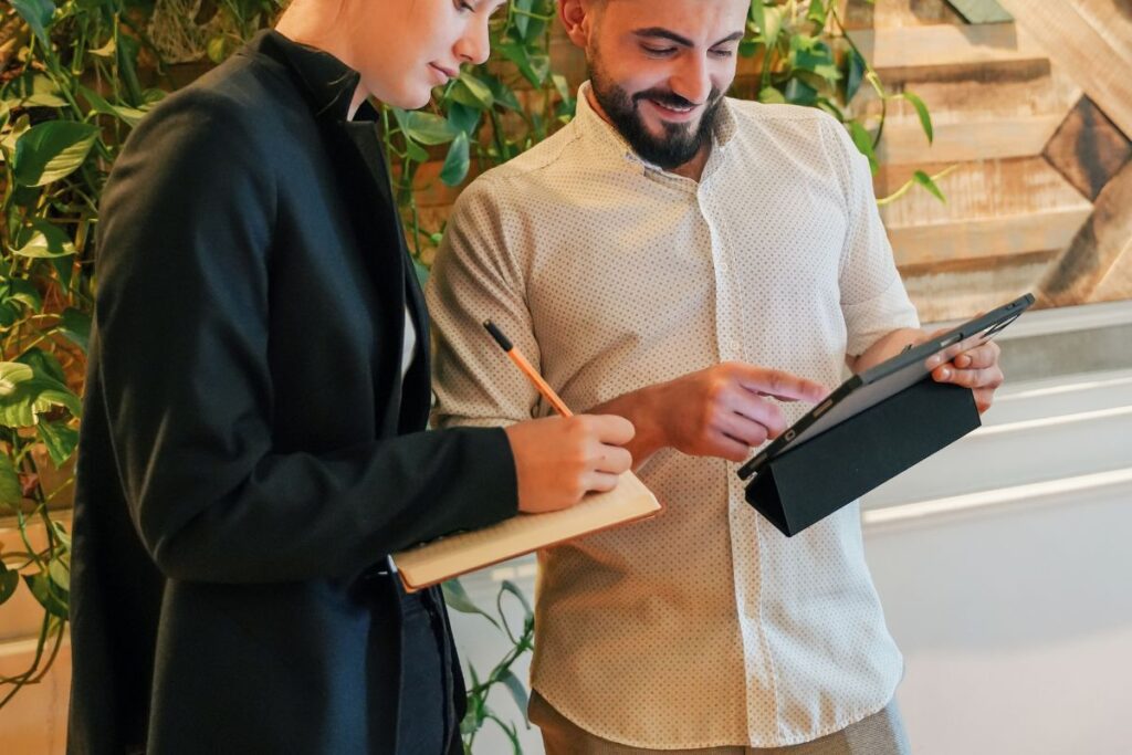 A woman writes using an eco-friendly pencil. Her male colleague scrolls on his iPad next to her.