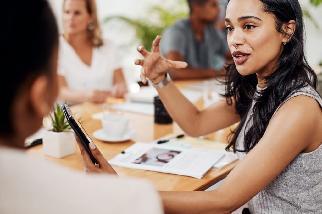 A woman collaborates with a colleague in a meeting. She is holding an iPad in one hand, and holding her other hand in the air as she speaks.