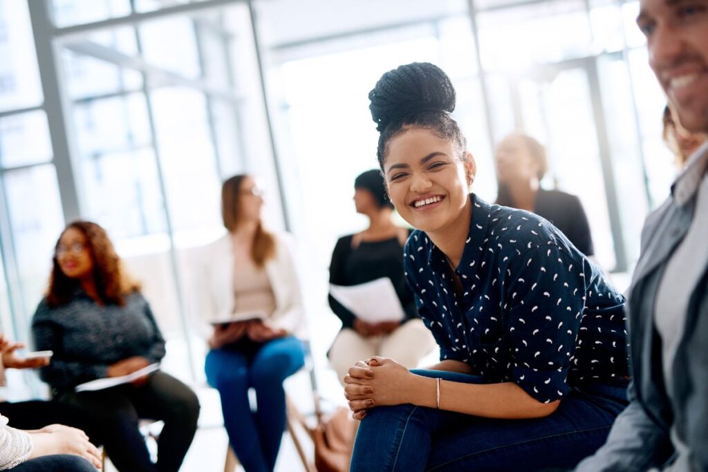 A woman sits in a meeting and smiles. She is wearing a bun and jeans.