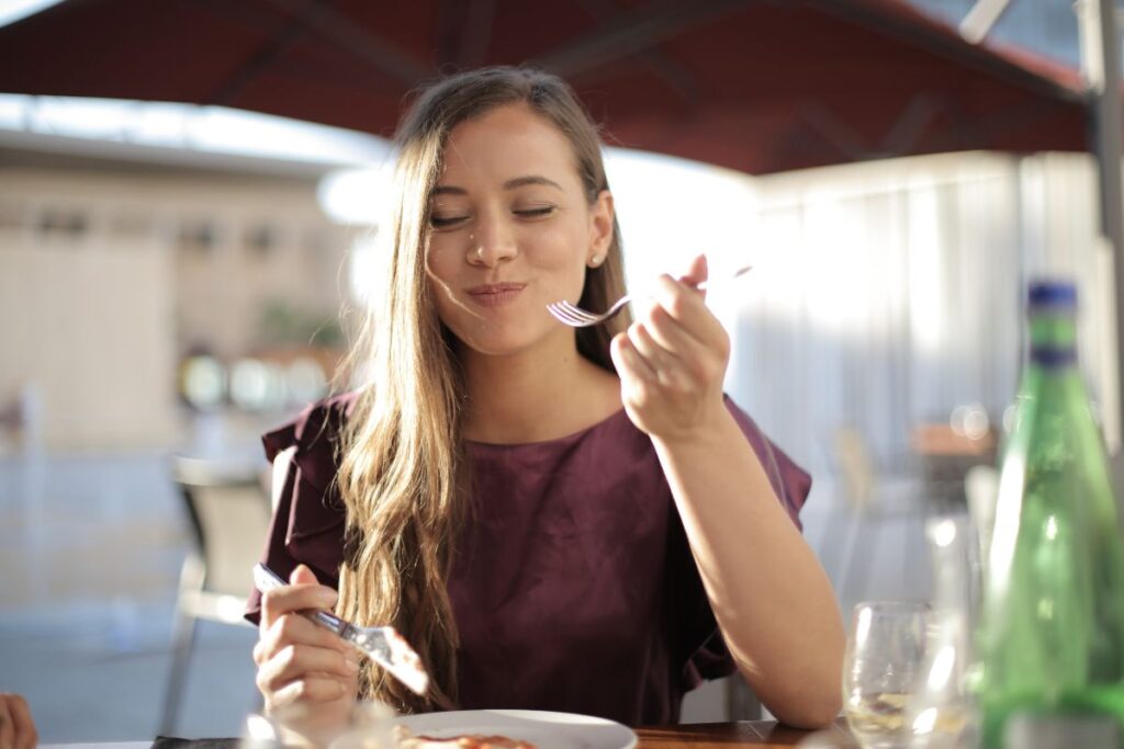 A woman eats a meal in the sun and smiles. She holds a fork to her mouth and has her eyes closed.