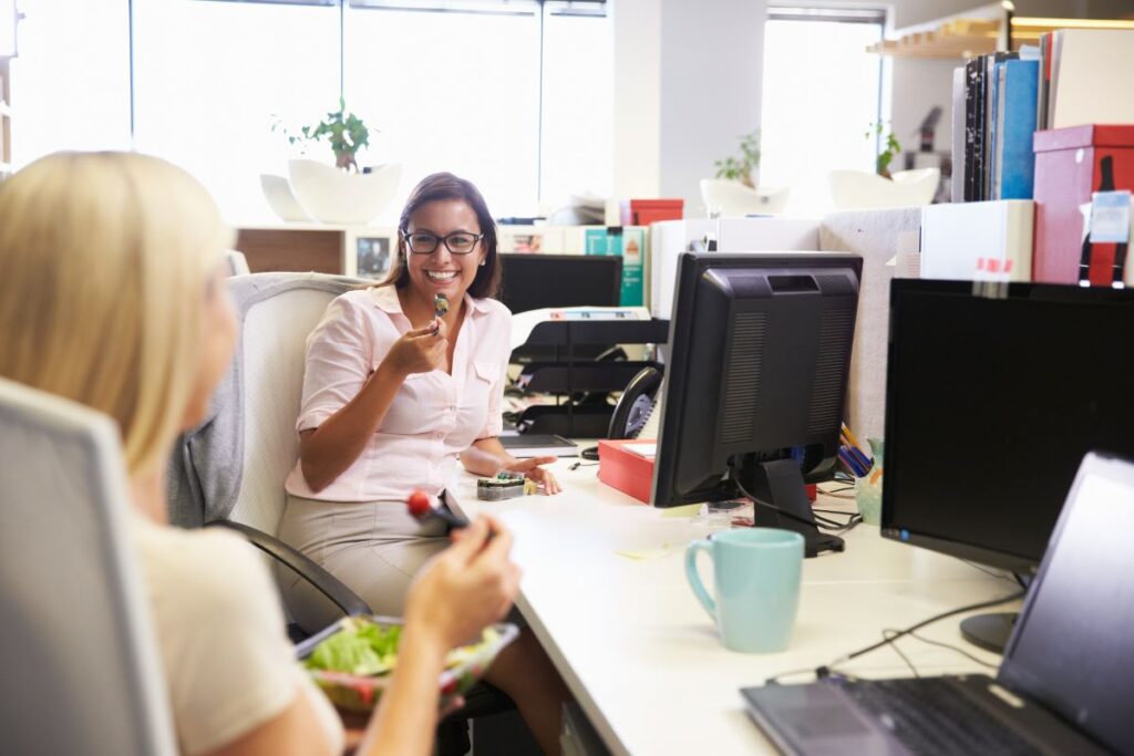 Two women sit at their desks in the office and eat salads and vegan sushi. They are smiling.