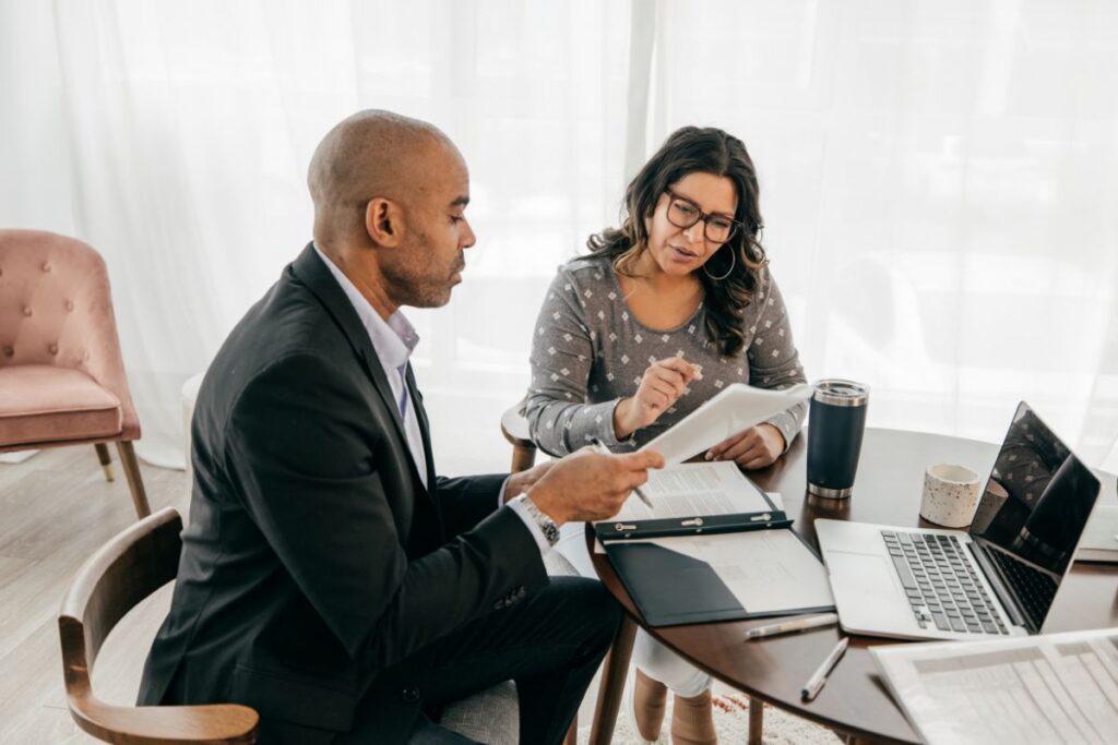 Two co-workers sit together in a meeting to work out a performance management process. There is a laptop on the table and lots of notes.