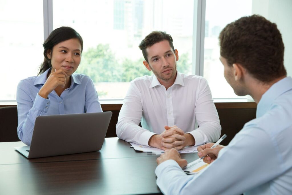 three coworkers sit together in a meeting. They all wear formal workwear. There is a laptop on the table.