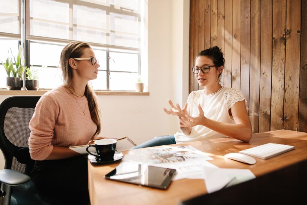 Two women sit in an office and discuss performance management. The desk is full of notes, as well as an iPad and a coffee cup.