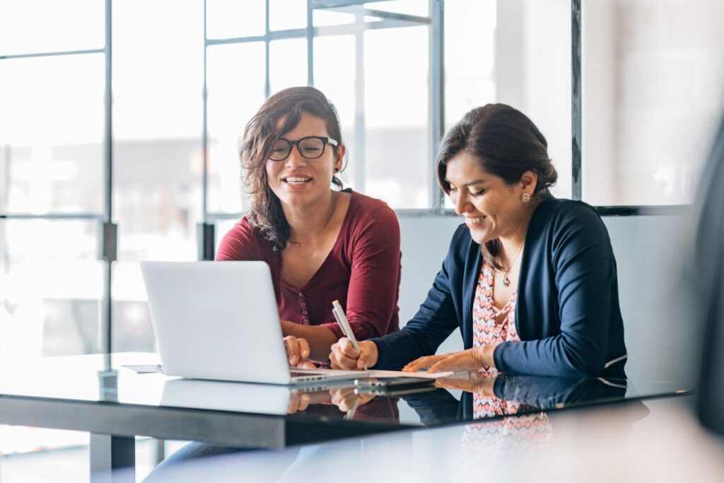 Two colleagues sit together and work on a laptop. They are both smiling. The woman on the right writes down notes, whilst the woman on the left scrolls on the laptop.