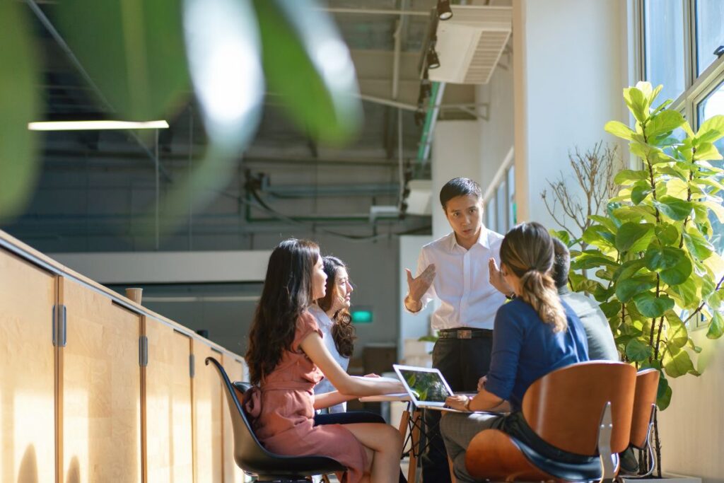 A sales meeting takes place in an office. One man is stood up at the head of the table. 