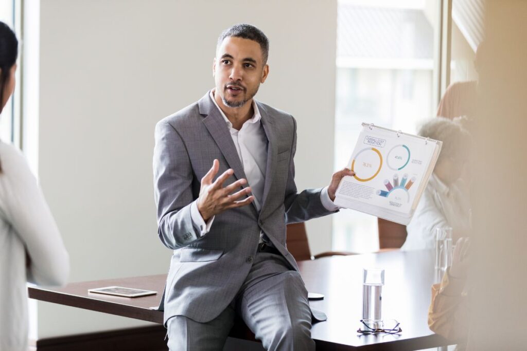 A man presents data in a sales meeting. He is wearing a suit and perching on the side of a table. 