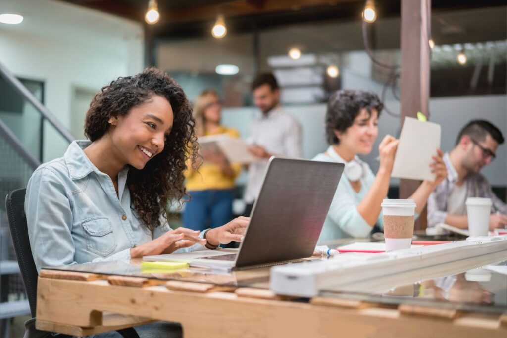 A woman sits at her desk in a co-working space and smiles at her laptop.