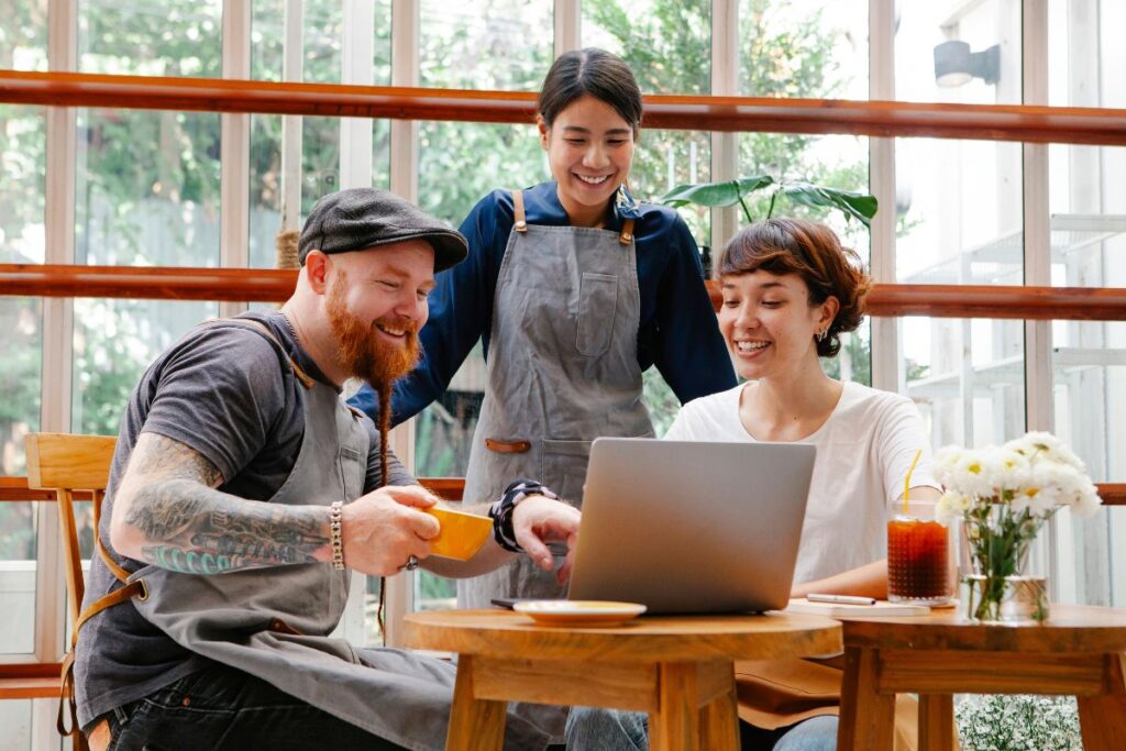 A group of three employees work together in a café. They are all smiling and chatting whilst working through a project on their laptop.