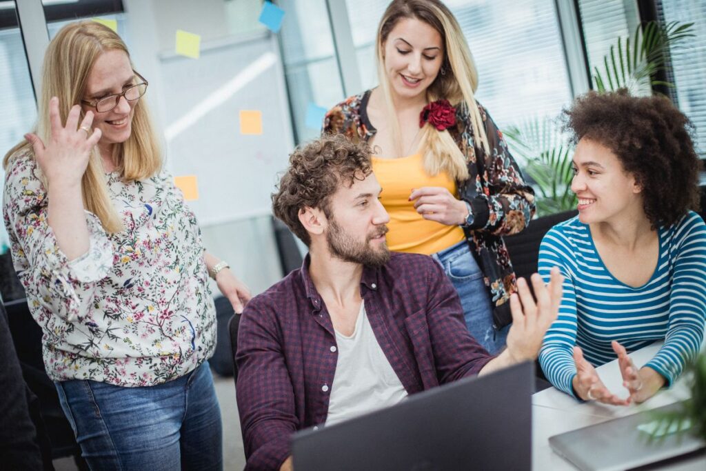 A group of four co-workers collaborate on a project whilst at their desks. They are all smiling as they talk.
