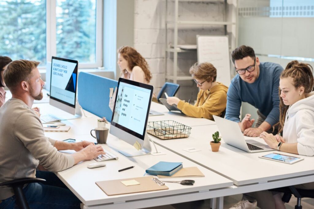 A group of employees sit in their co-working space. One man offers support to one woman, while the rest of the group focus on their work.