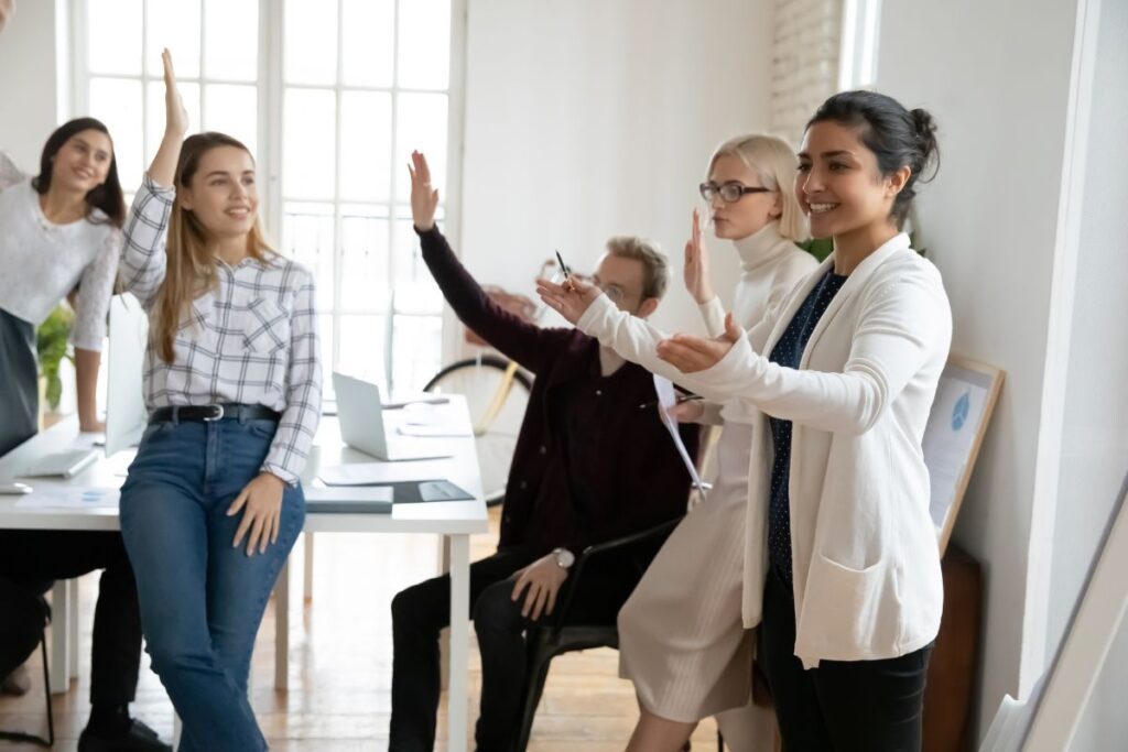 A group of employees listen to a presentation. They are smiling and holding their hands in the air in participation