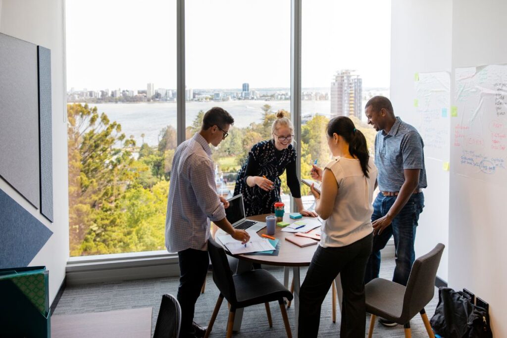 A team of four employees work together in a breakout room. They are all stood up around a table. There is a view of a city in the background.