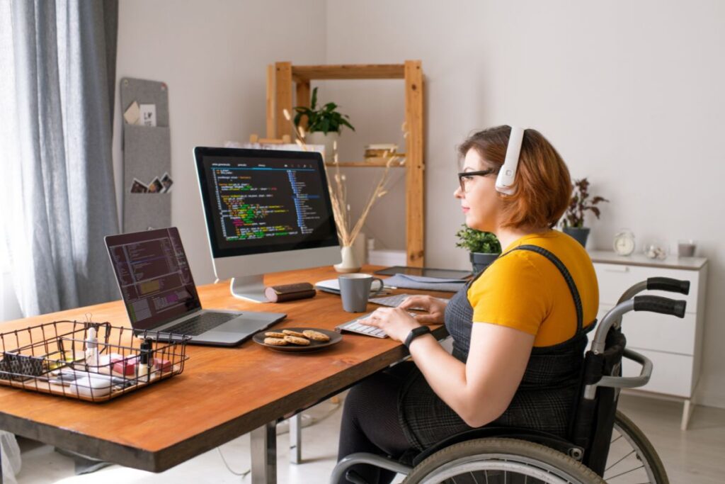 A woman in a wheelchair works from home. She is sat at her desk with a laptop and a monitor in front of her. She is coding.