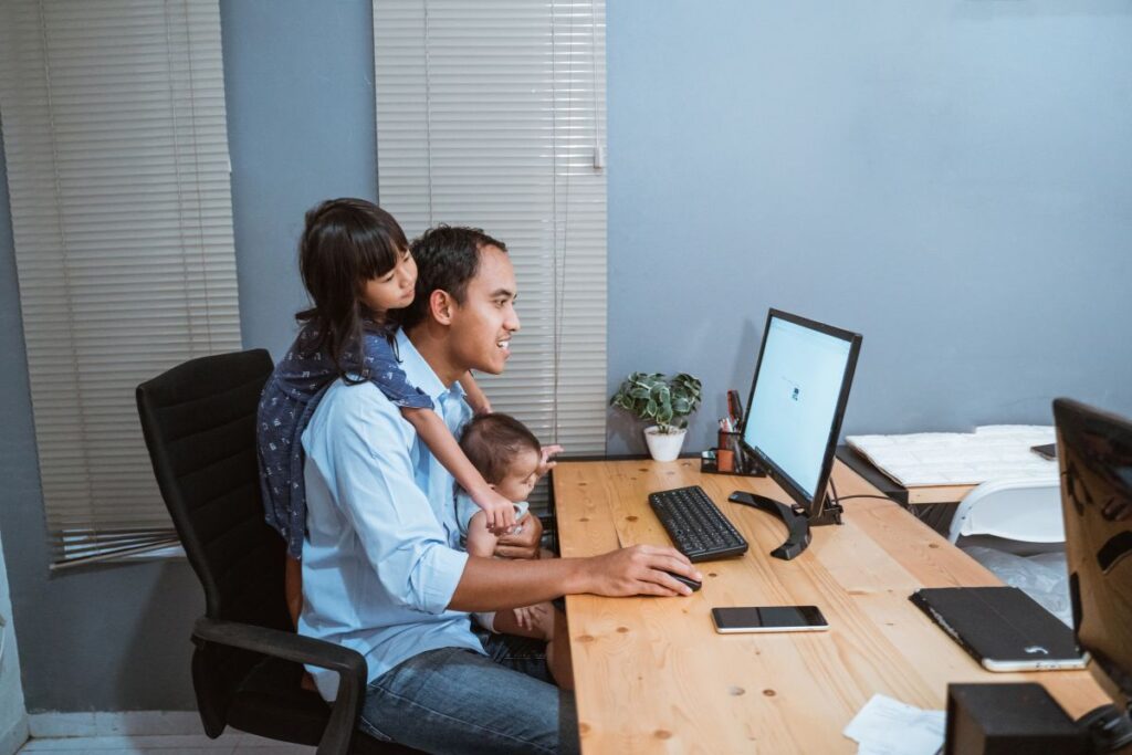 A dad works from home at his desk. He holds his baby on his lap and his young daughter holds onto his shoulders. He is smiling as he works on his computer.
