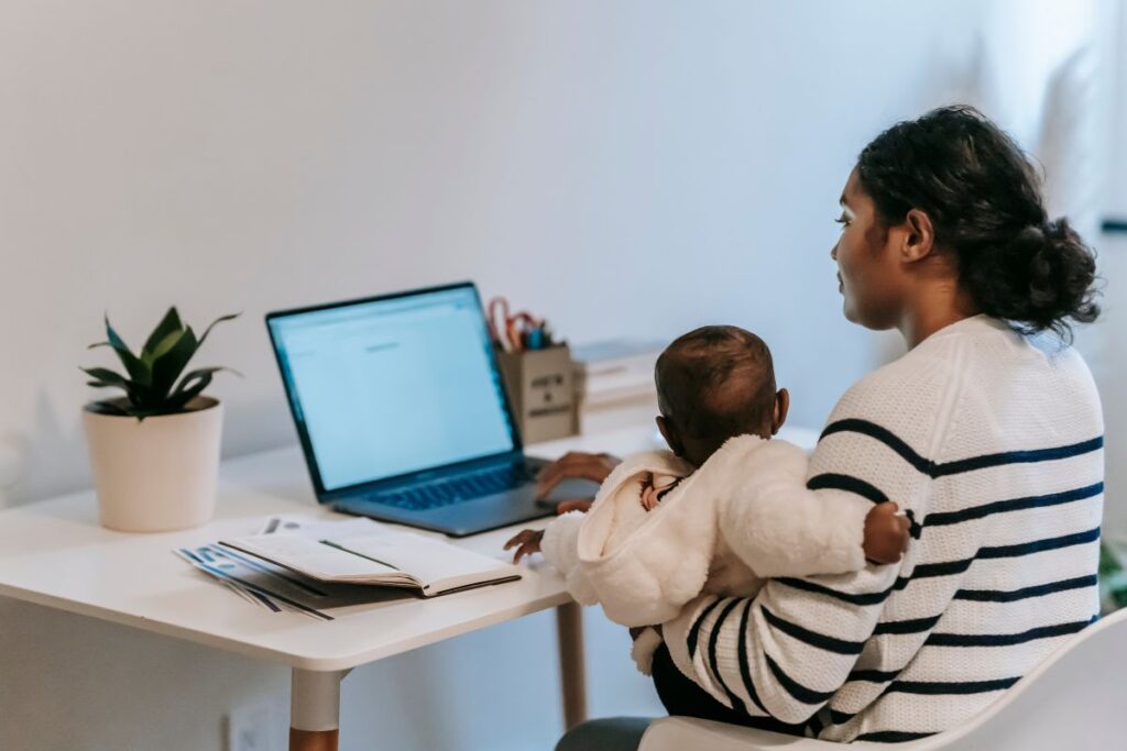 A new mother sits at her desk and works from home. She holds a baby in her arms and works on her laptop.