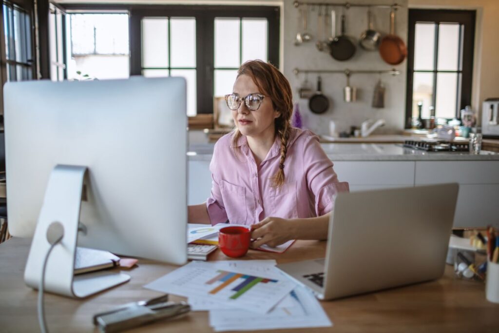 A woman sits at her desk in her kitchen and works from home. She works at her laptop. There are sheets of graphs on the table. She is holding a mug.