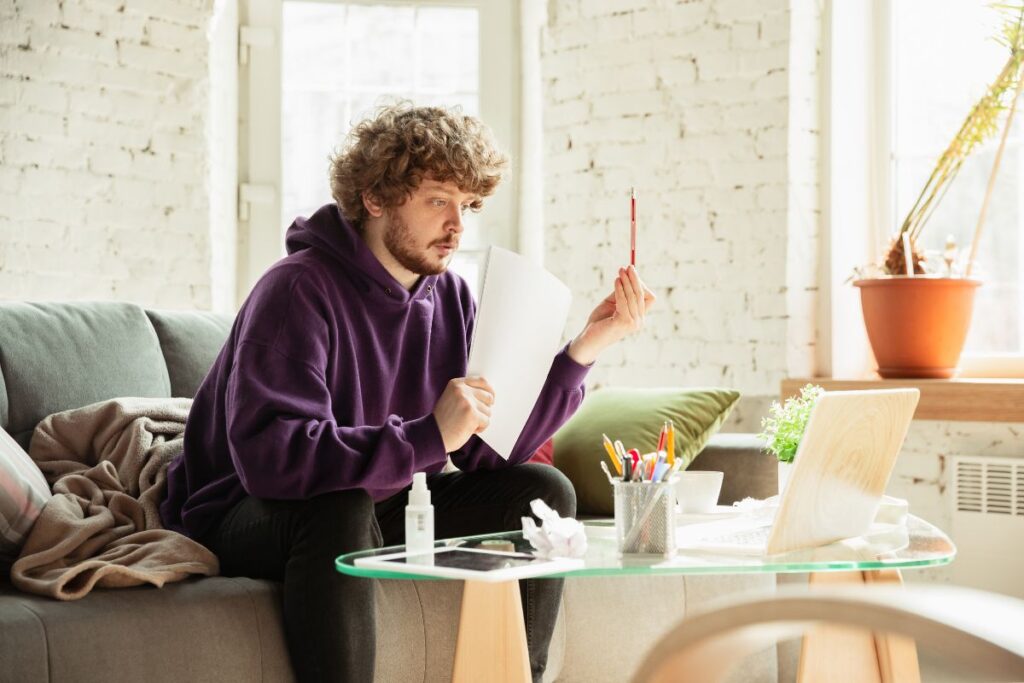 A man works from home. He sits on his sofa, holding sheets of paper and a pen.