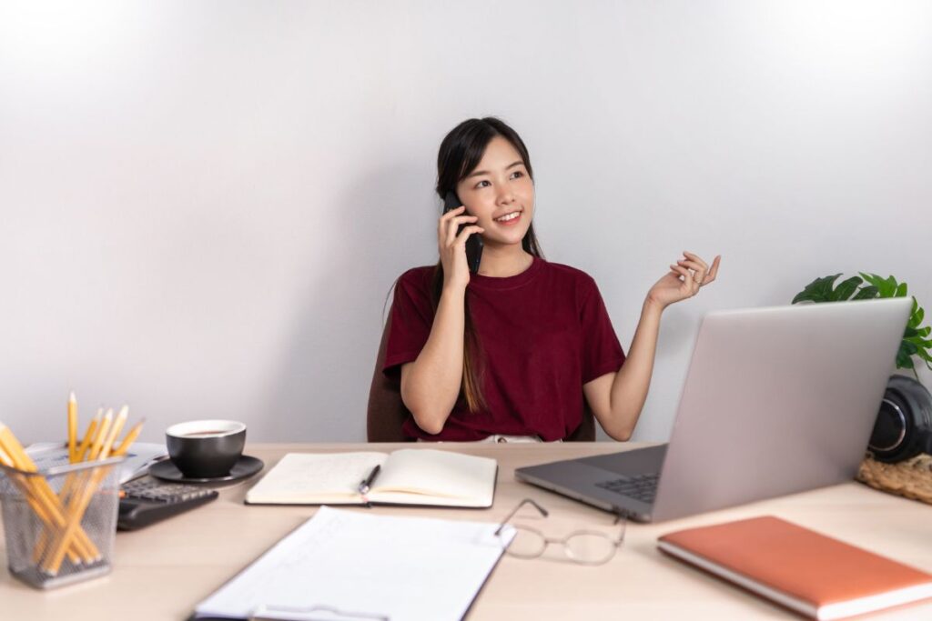 A woman sits at home and works at her desk. There is a laptop on her desk alongside notebooks and a pair of glasses. She smiles as she talks on the phone.