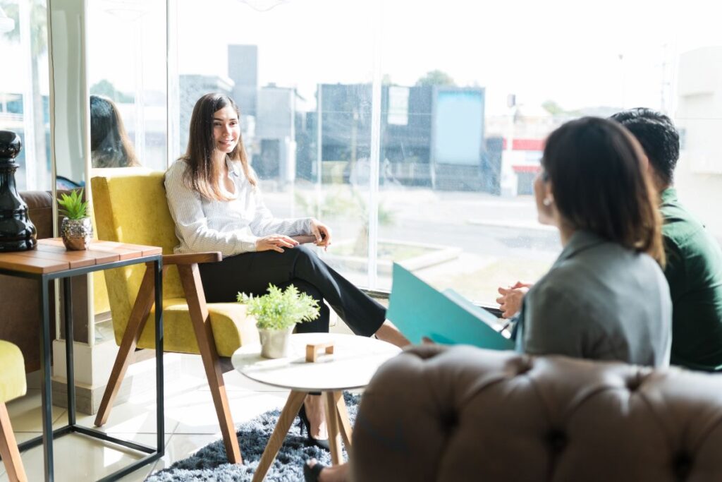 Three people sit by a window in a workplace and smile. One of them holds a clip board. They are all dressed in formal workwear.