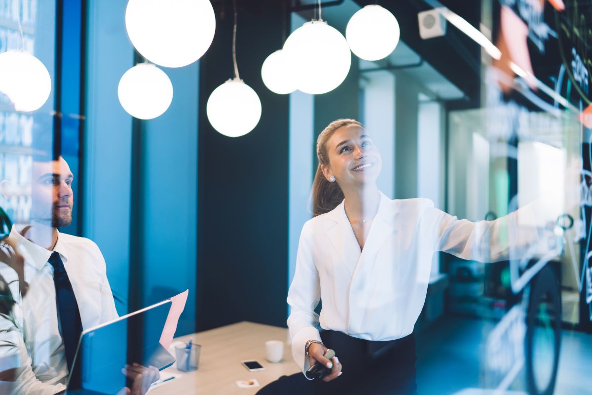 A woman smiles as she presents in front of a team. The photo is taken through the window of a boardroom. The lights reflect off the glass.